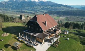 A charming building with a large garden and outdoor terrace. In the background, snow-capped mountains and a green landscape can be seen.