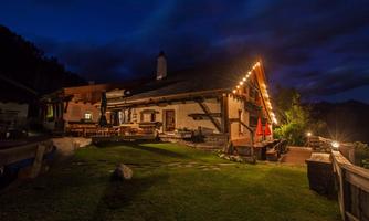 A cozy alpine hut with warm light, illuminated at night. On the meadow, there are tables and chairs, surrounded by mountains and a clear sky.