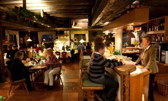 A cozy restaurant with wooden decor and warm lighting. Guests are relaxed and chatting at the table and at the bar.