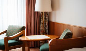 A cozy seating area with two green chairs and a small table. The windows are covered with light curtains, and the atmosphere feels inviting.
