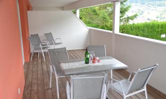A cozy terrace with a table and six chairs. On the table, there is a bottle of wine and some glasses.