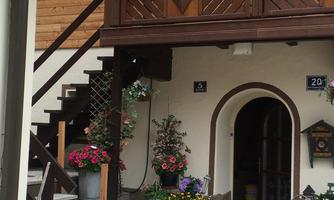 An inviting entrance to a house with wooden cladding and blooming plants. The staircase leads to a balcony that is also adorned with flowers.