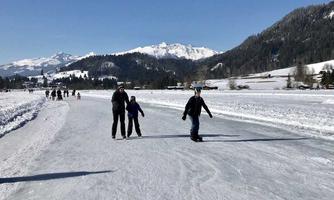 A winter ice rink with people ice skating. In the background, there are snowy mountains and a clear blue sky.