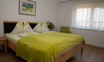 A modern bedroom with a large bed and a green blanket. A picture hangs on the wall, and the room has a pleasant, bright light.