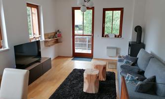 A modern living room with a gray sofa, a large television, and two wooden tables. The room has large windows that provide plenty of daylight and a view of nature.