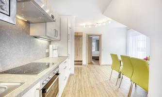 A modern kitchen with shiny surfaces and modern appliances. In the background, a dining area with green chairs can be seen.