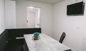 A simple dining room with a table, a black bench, and a television on the wall. The room is bright and minimally designed.