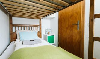 A simple bed in a small room with a wooden frame and a cozy blanket. On the wall, there is a door and a small shelf.