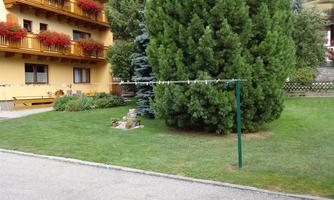 A well-maintained garden with a large green tree and a clothes rack. In the background, a light yellow building with balconies and flowers can be seen.
