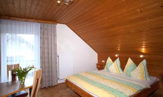 A cozy bedroom with wooden walls and a large window. The bed is decorated with colorful pillows and a small table stands beside it.