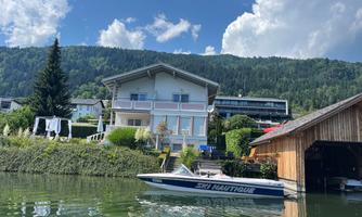 A modern house by the water with a boat in the foreground. In the background, hills and a blue sky can be seen.