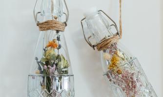 Two decorative glass bottles hang on a wall and are filled with dried floral decorations. They are wrapped with jute twine, adding a rustic touch.