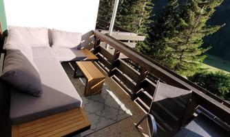 A cozy balcony view with a lounge chair and a small table. In the background, tall fir trees and a beautiful mountain landscape can be seen.