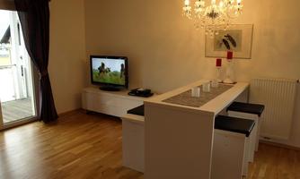 A modern living room with a table and chairs. A TV is placed on a cabinet and an elegant ceiling lamp hangs from the ceiling.