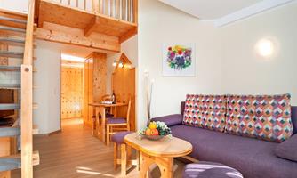 A bright living room with a cozy couch and colorful pillows. In the background, a staircase and a dining area are visible.