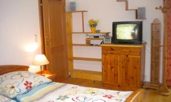 A cozy bedroom with a wooden bed and colorful bedding. A television is on a shelf in the corner, and a lamp provides soft light.