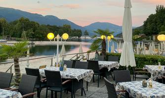 A charming restaurant with a view of a lake and mountains in the background. Tables are set with blue tablecloths and umbrellas are ready.