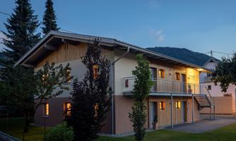 A modern building with wood cladding and warm lights in the windows. Surrounded by trees and a quiet environment.