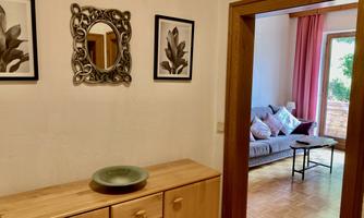 A bright living room with a gray couch and colorful cushions is visible through an open passageway. Two pictures of plants hang on the wall, and a wooden cabinet stands in the foreground.