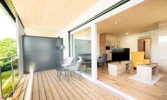A modern balcony with a wooden floor and comfortable seating. In the background, a bright, stylish living and dining room can be seen.