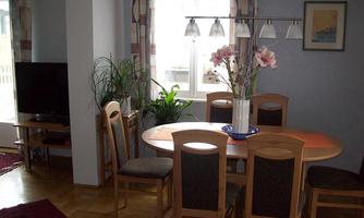 A cozy dining area with a round table and six chairs. The windows let in plenty of light and there is a plant as well as a flower vase on the table.