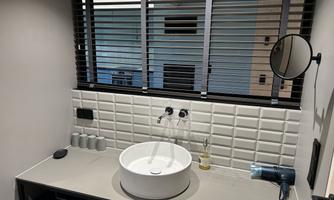 A modern bathroom with a round sink and a hairdryer. The windows are covered with horizontal blinds and the walls are tiled in white.