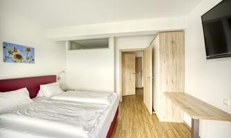 A modern bedroom with a double bed and a wooden floor. A picture of sunflowers hangs on the wall and a television is mounted.