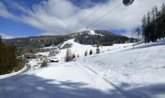 A winter landscape with snow-covered hills and a cable car in the background. The sun is shining and the sky is partly cloudy.