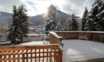 A snowy landscape with green fir trees and mountains in the background. The sun is shining on a wooden terrace.