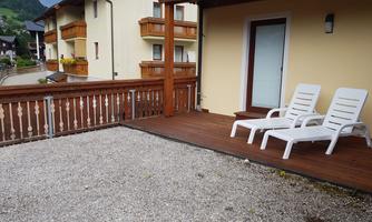 A terrace with wooden decking, featuring two loungers. In the background, several buildings and a green landscape can be seen.