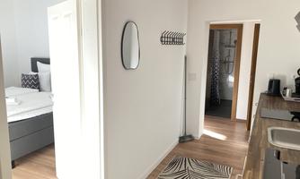 A bright hallway with wooden flooring that leads to a bedroom and a bathroom. A round mirror and a towel rack are hanging on the wall.