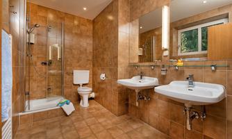 A modern bathroom with brown tiles and a large mirror. There is a shower cabin, two sinks, and a toilet.