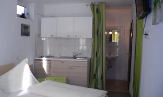 A small room with a bed and a kitchenette. In the background, there is a bathroom and a television on the wall.