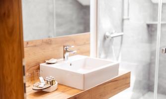 A modern bathroom with a simple sink and wooden shelf. On the shelf are placed care products and glasses.