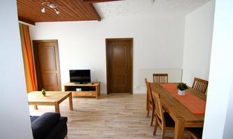 A bright living room with a sofa, a table, and a television. The adjacent dining area has a large wooden table with chairs.