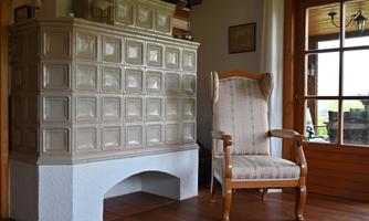 A cozy room with a tiled stove and a comfortable armchair. The window lets in plenty of daylight.
