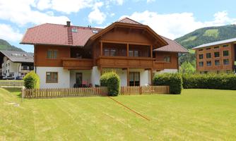 A large, traditional wooden house with a balcony and a well-maintained garden. In the background, gentle hills and a blue sky can be seen.