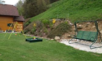 A cozy garden with a swing and a bench. In the background, there is a wooden house and a well-maintained slope with flowers.