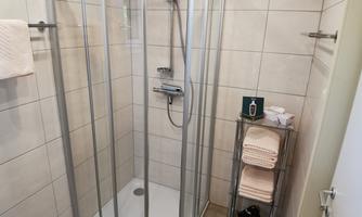 A modern shower with a glass partition is in a bright bathroom. Next to the shower are towels and a shelf with additional towels.