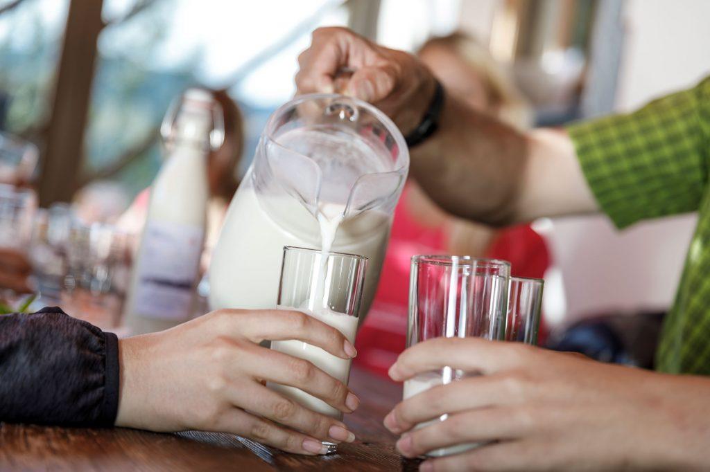 Milch wird aus Glaskrug in Gläser auf einem Holztisch gegossen, im Hintergrund Flasche
