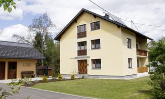 A modern, two-story house in yellow color with wooden balustrades. It is located in a quiet environment with lawn and trees.