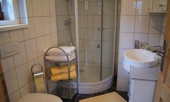 A small bathroom with a shower, sink, and toilet paper holder. Towels are neatly stacked on a shelf.