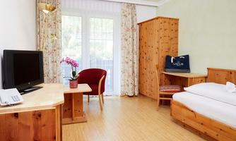 A cozy room with wooden furniture and a television.  
There is a small table with a flower and a chair next to a window.