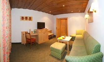 A cozy room with a wooden beam ceiling, green sofas, and a table. There is a TV and a small workspace.