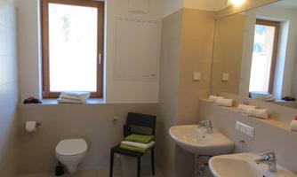 A modern bathroom with bright tiles and a large window. There are two sinks, a toilet, and a chair with towels.
