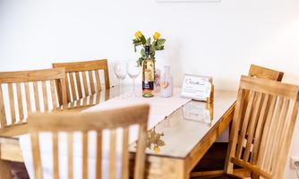 A beautiful wooden dining table with chairs and a tablecloth. On the table are a bottle of wine, glasses, and a vase with fresh flowers.
