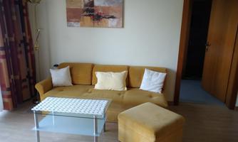 A cozy living room with a yellow sofa and a glass table. The room is bright and inviting, with a piece of artwork on the wall.