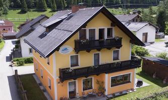 A yellow house with balconies and solar panels, surrounded by a green garden. In the background, there are forests and mountains visible.