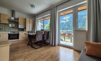 A bright and modern apartment with an open kitchen and dining area. Large windows offer a beautiful view of nature.