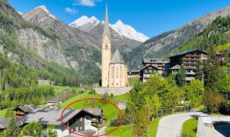 A picturesque mountain landscape featuring a tall church tower and snow-capped peaks in the background. In the foreground, traditional alpine houses and green meadows can be seen.